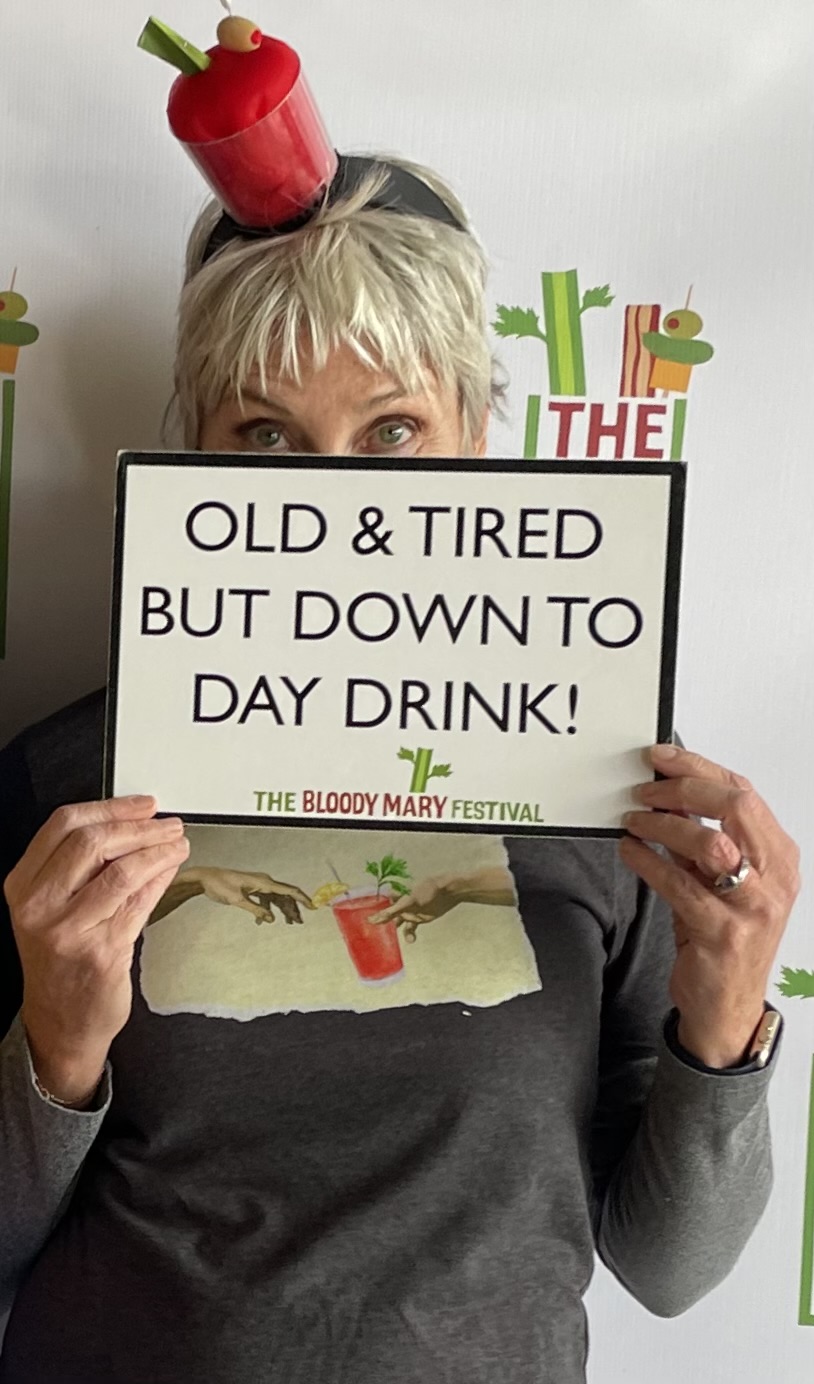 Person wearing a red cup hat holds a sign reading “Old & tired but down to day drink!”
