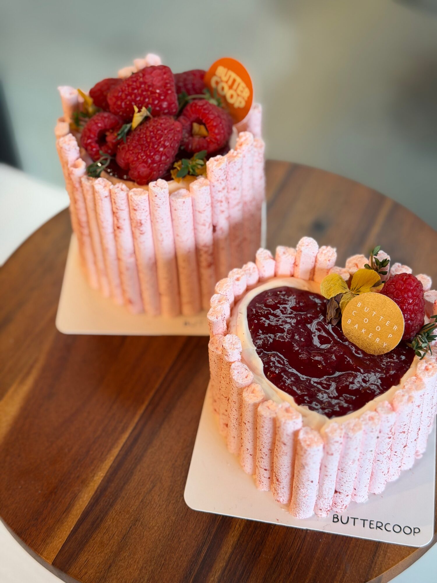 Two pink-frosted cakes topped with berries, fruit, and chocolate sit on a wooden table.