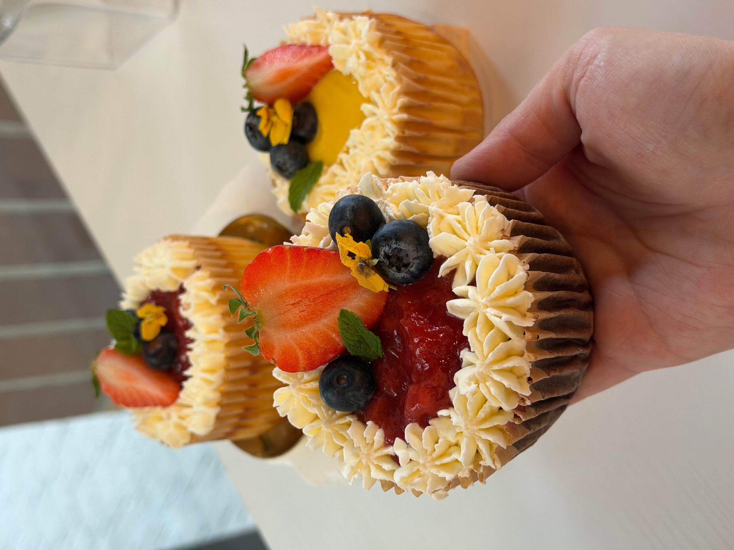 A hand holds three small decorated cakes topped with piped cream, strawberries, blueberries, and dark fruit filling.