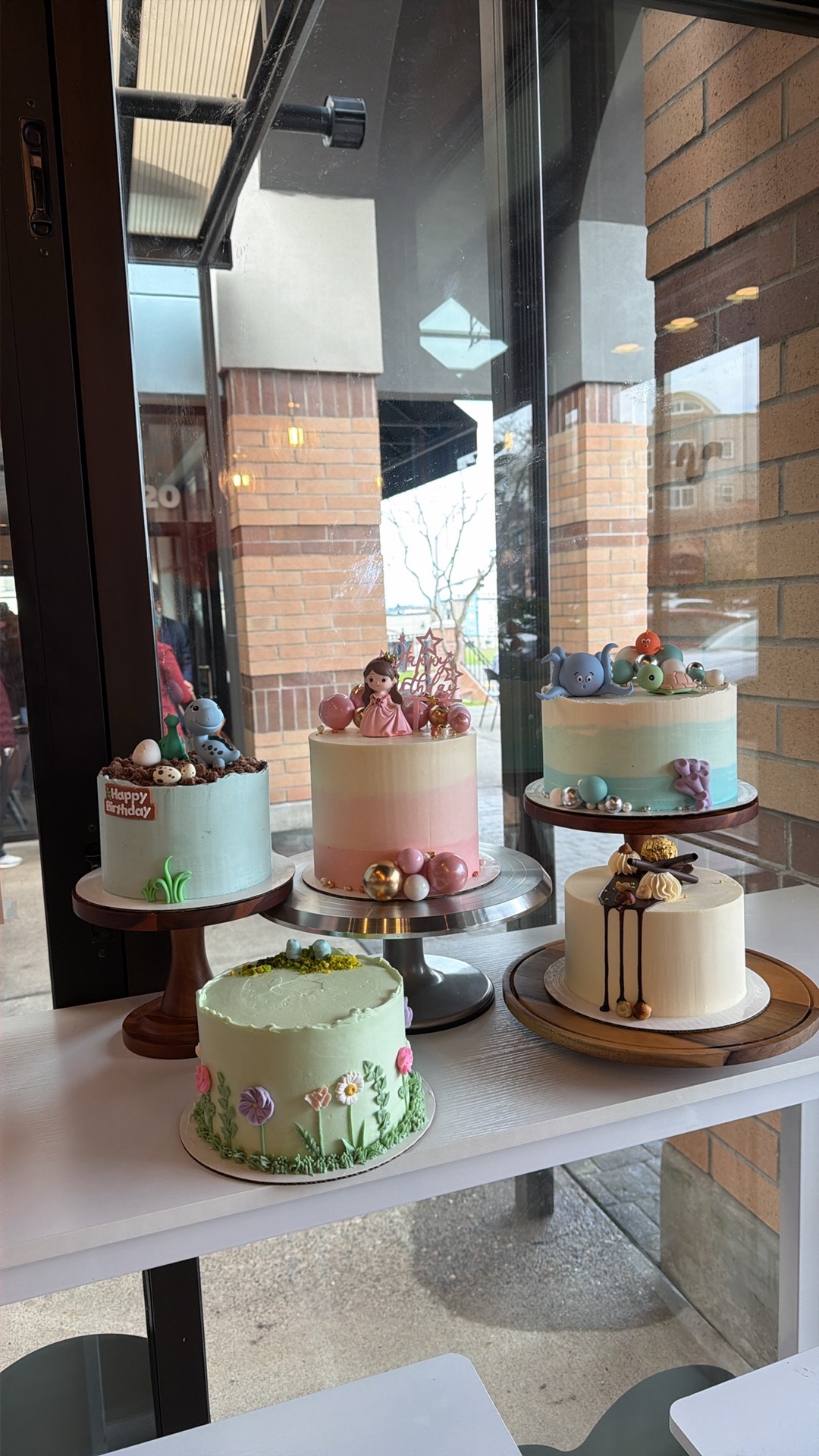 Several decorated cakes sit on round stands inside a glass-walled bakery or café.