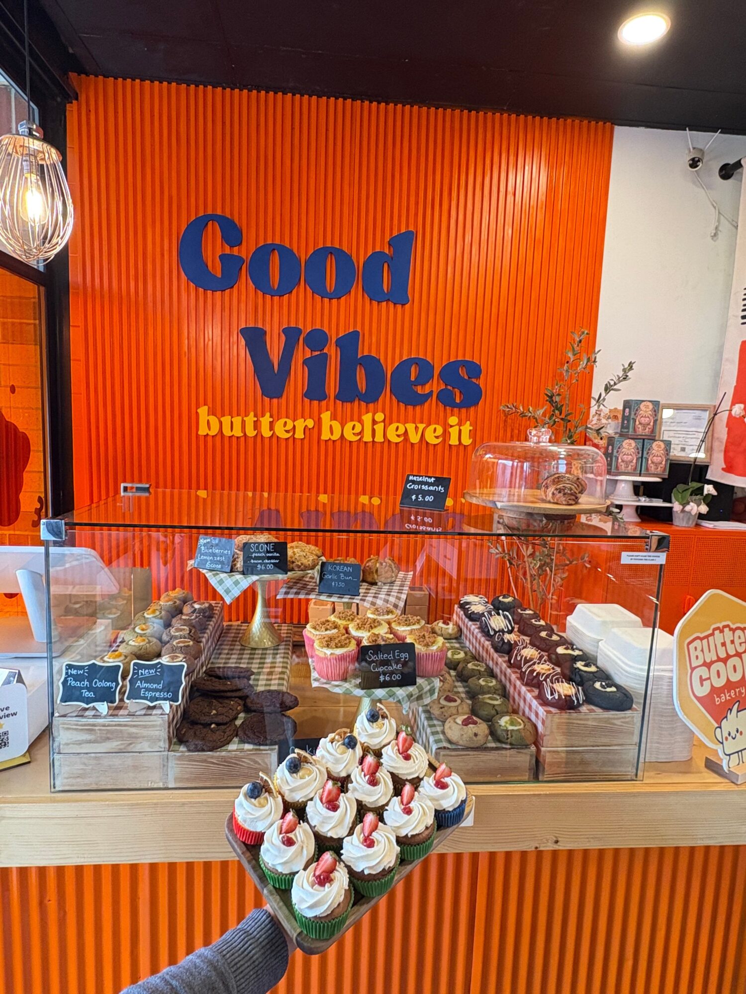 Glass display case with rows of baked goods in front of an orange wall reading “Good Vibes, Butter Believe It.”