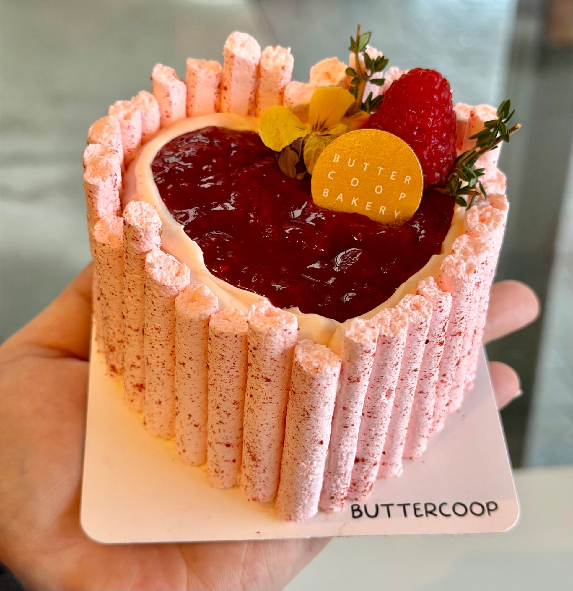 A heart-shaped cake with pink piped edges, red filling, a strawberry, flowers, and a yellow butter cookie, held on a plate.