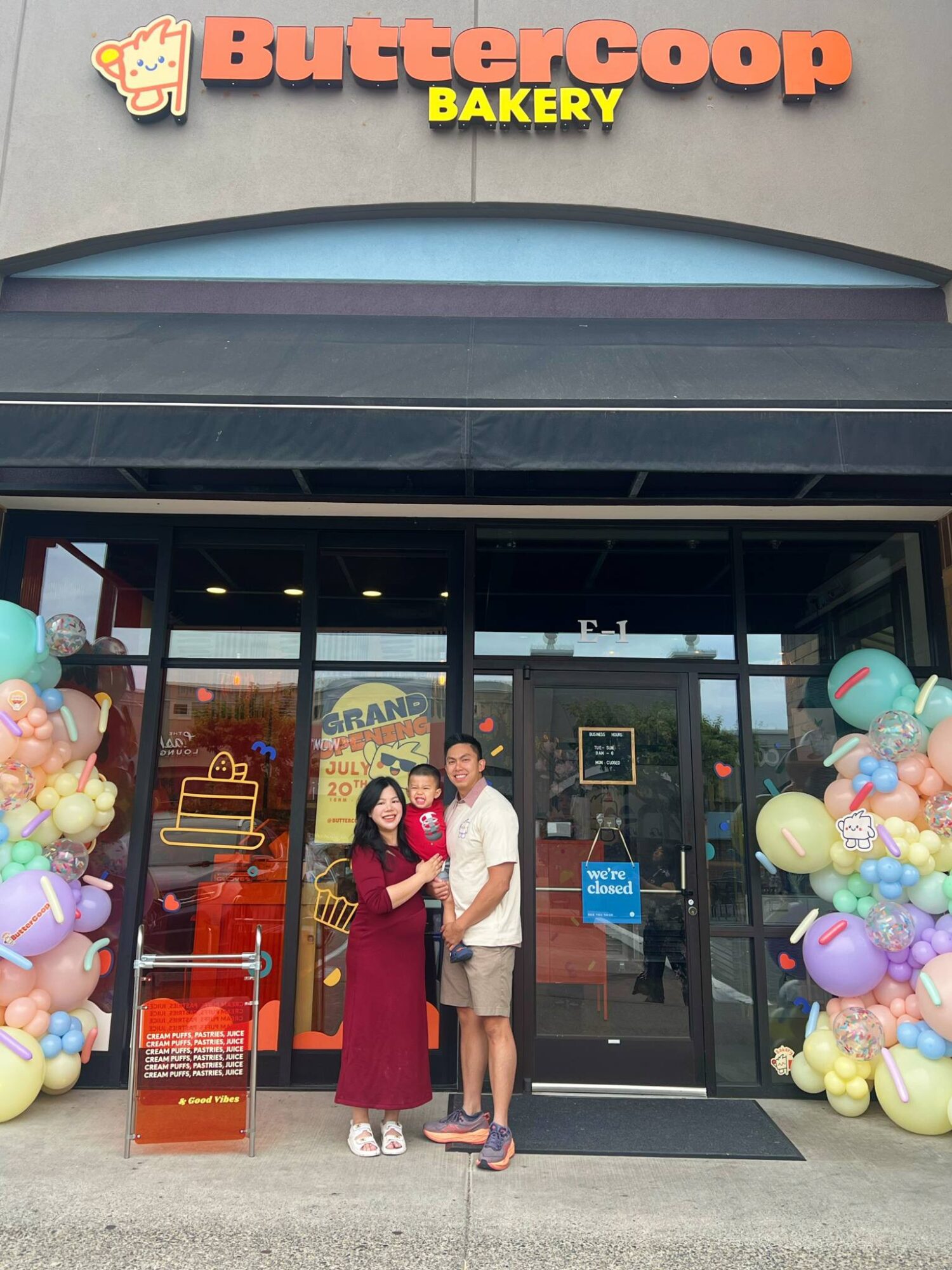 Two people stand at a bakery entrance decorated with colorful balloons and signs.