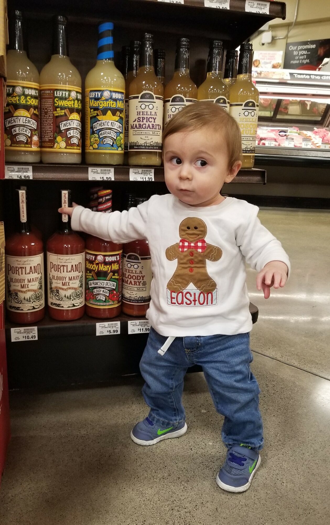 A toddler stands in a grocery aisle beside shelves of sauces, wearing a white shirt with a gingerbread figure.