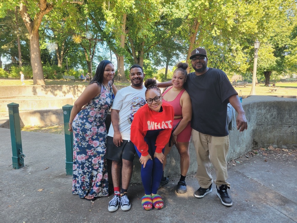 Six people pose together beside a low wall in a tree-lined park.