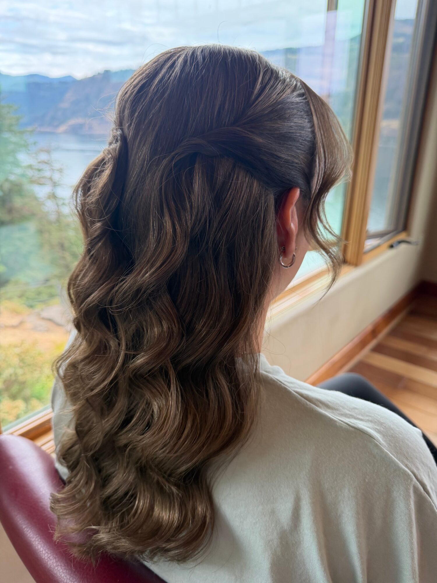 Person with long wavy hair seated beside a window overlooking water and mountains.