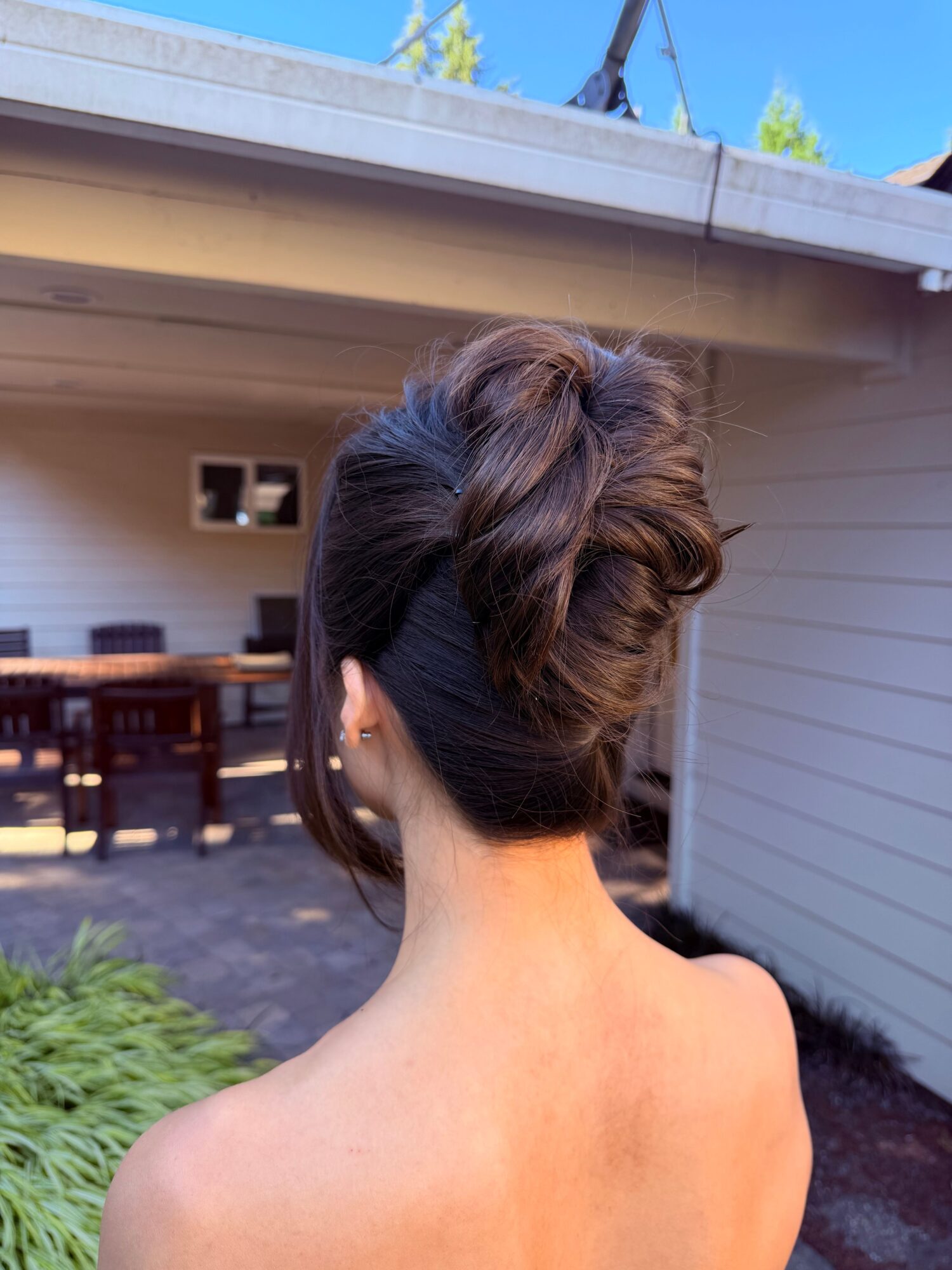 Person with dark hair in a bun standing beside a house, with a porch and greenery visible.