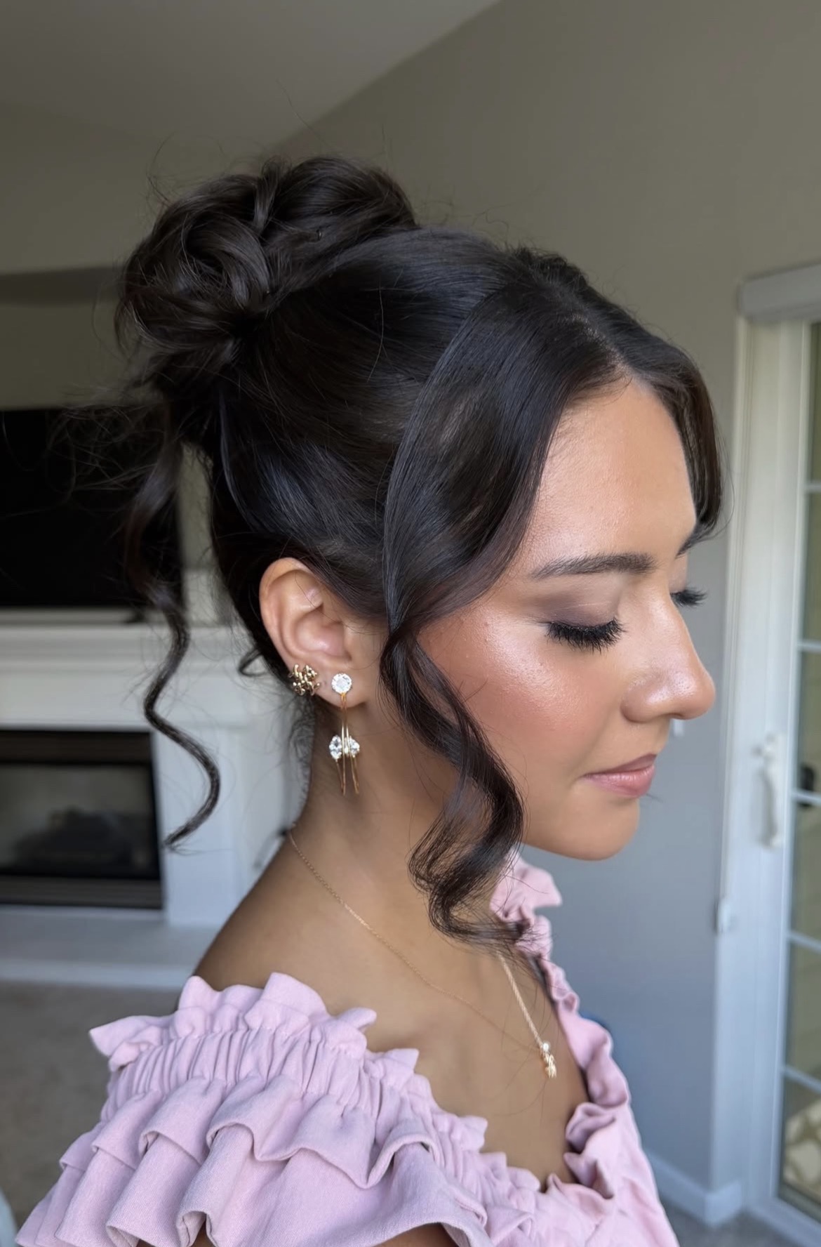 Person with dark hair in an updo, wearing earrings and a lilac ruffled top, shown in profile indoors.