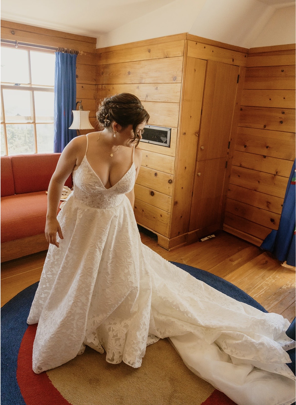 A person in a white lace gown stands in a wood-paneled room beside a window, with the train spread across the floor.