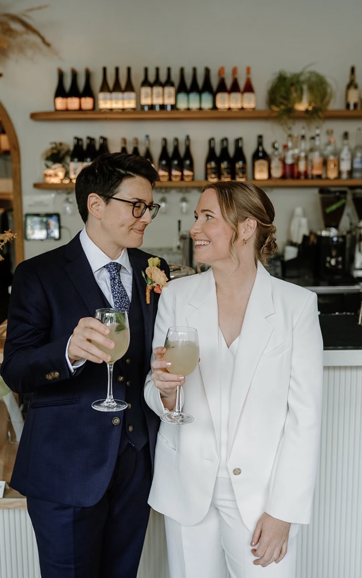 A person in a dark suit and a person in a white suit hold drinks and smile at each other in a bar.