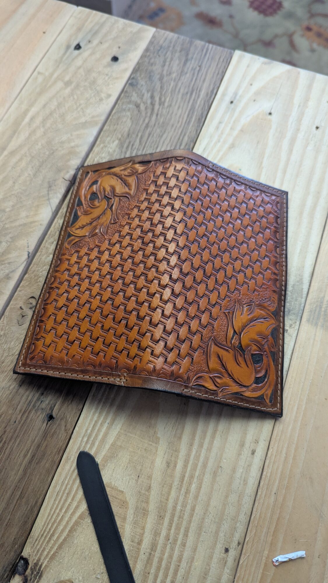 Brown embossed leather wallet resting on a light wooden plank floor, with a dark strap nearby.