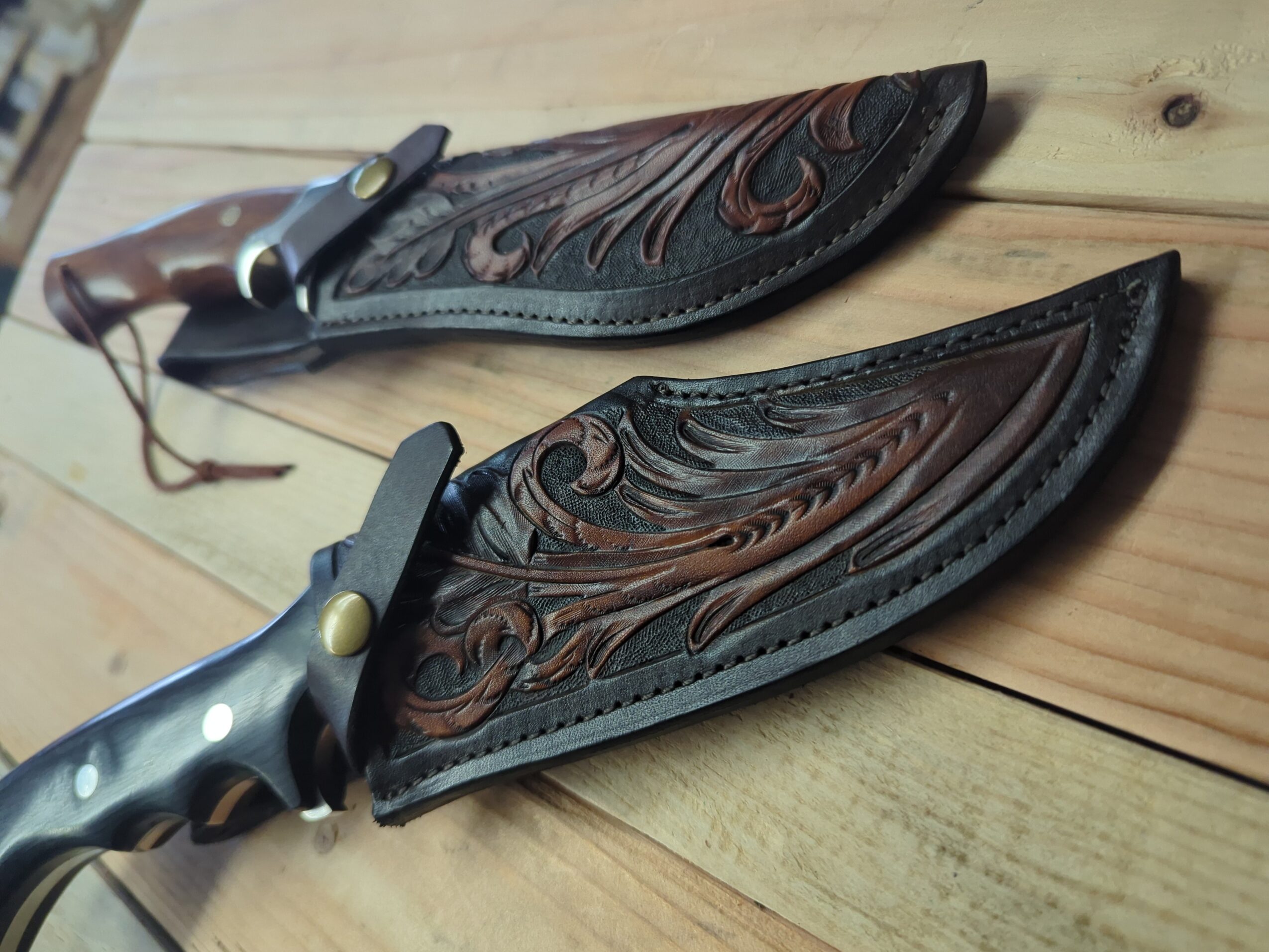 Two ornate leather knife sheaths with knives lie on a wooden surface.