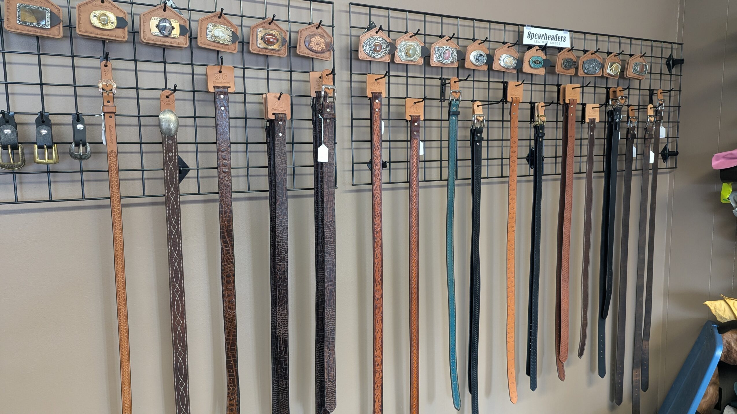 Belts hang vertically from a wall display grid, with small price tags above them.