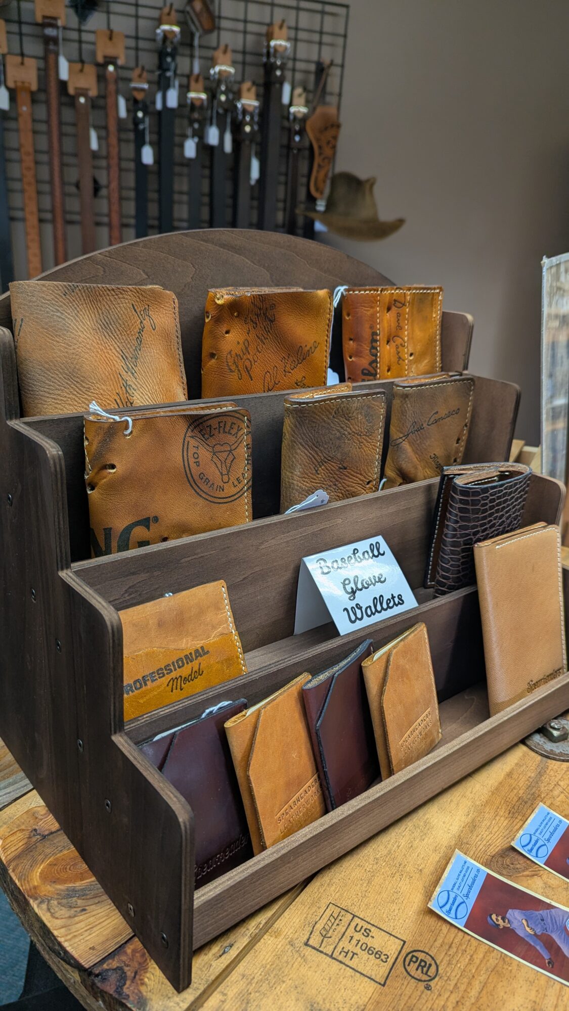 Wooden display rack holding leather goods, wallets, and small signs, with tools hanging on the wall behind it.