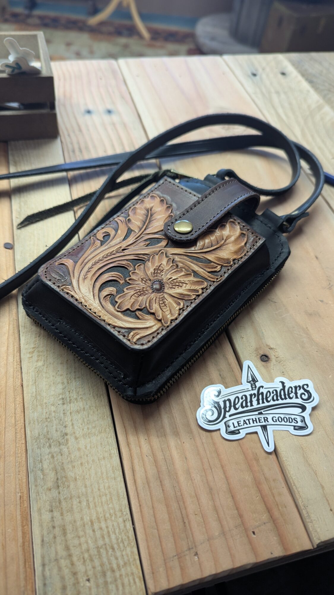 Ornate leather shoulder bag with a long strap on a wooden table beside a leather goods label.