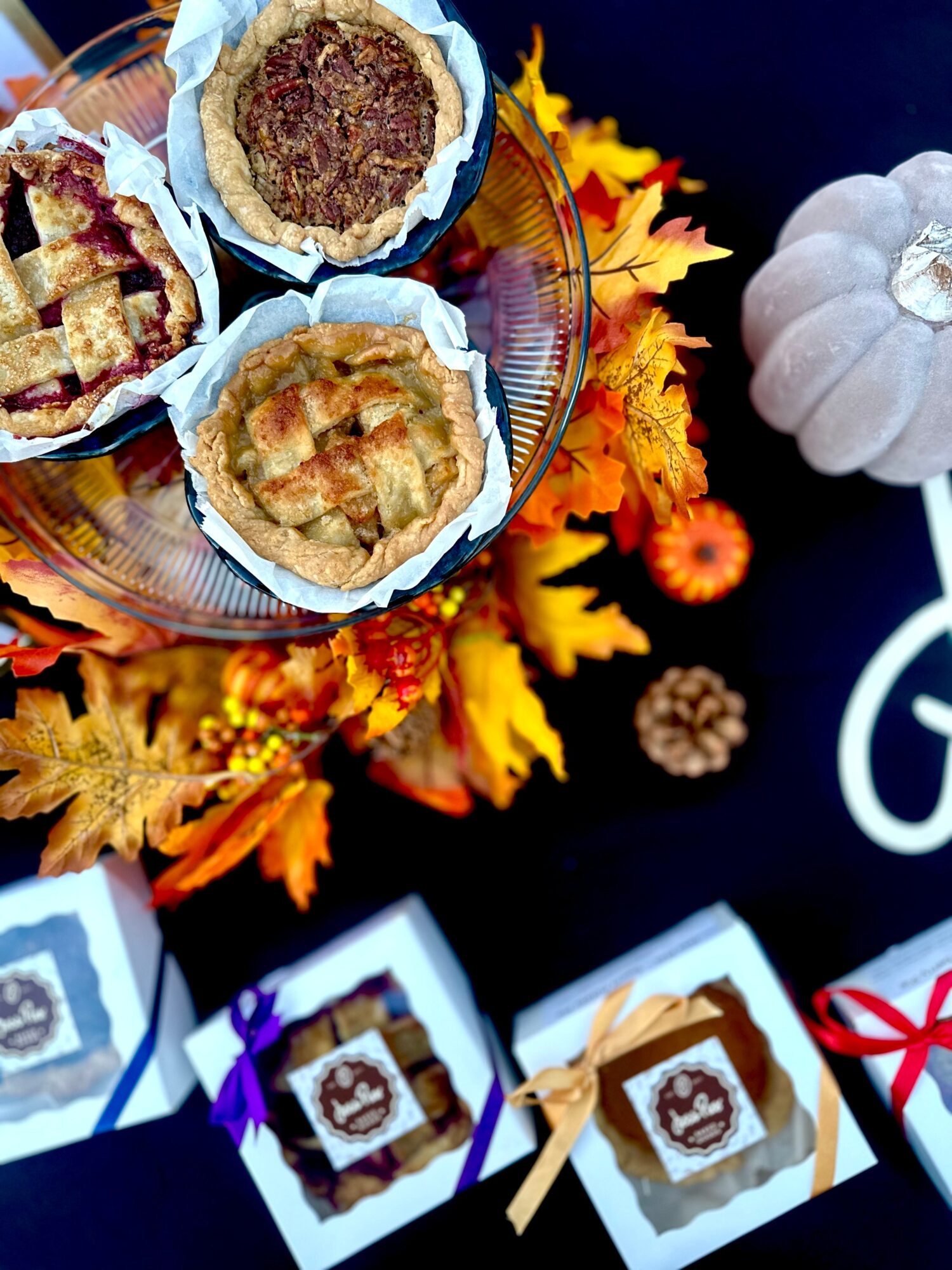 Three pies on a glass tray, autumn leaves, a white pumpkin, pinecone, and wrapped packages on a dark surface.