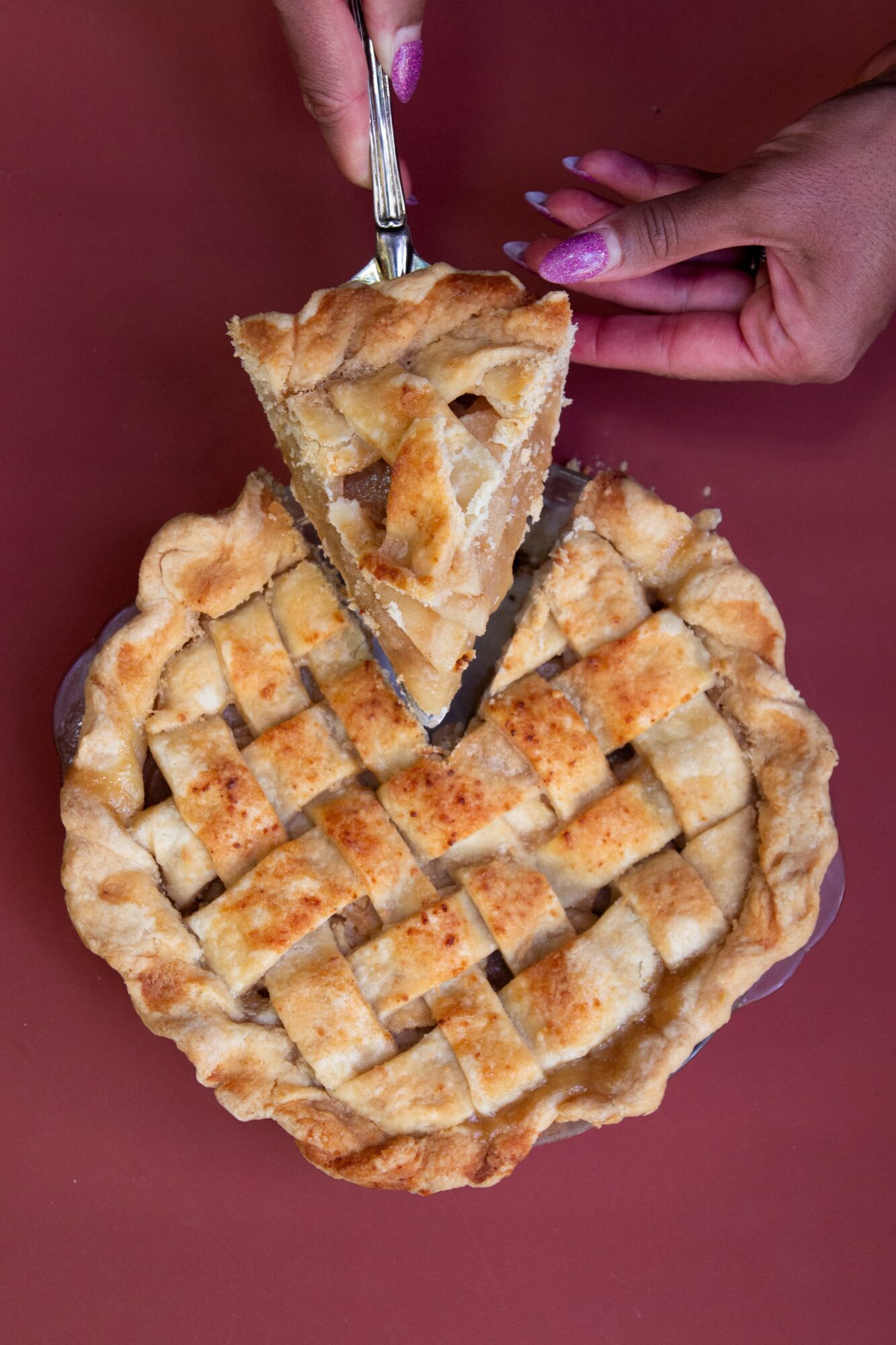 A lattice-topped pie is cut with a knife while a slice is lifted by hand.