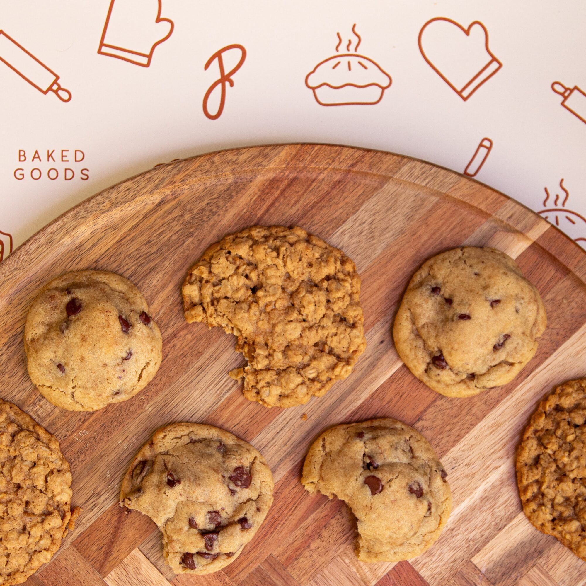 Cookies arranged on a wooden cutting board beside a wall decorated with baking illustrations.