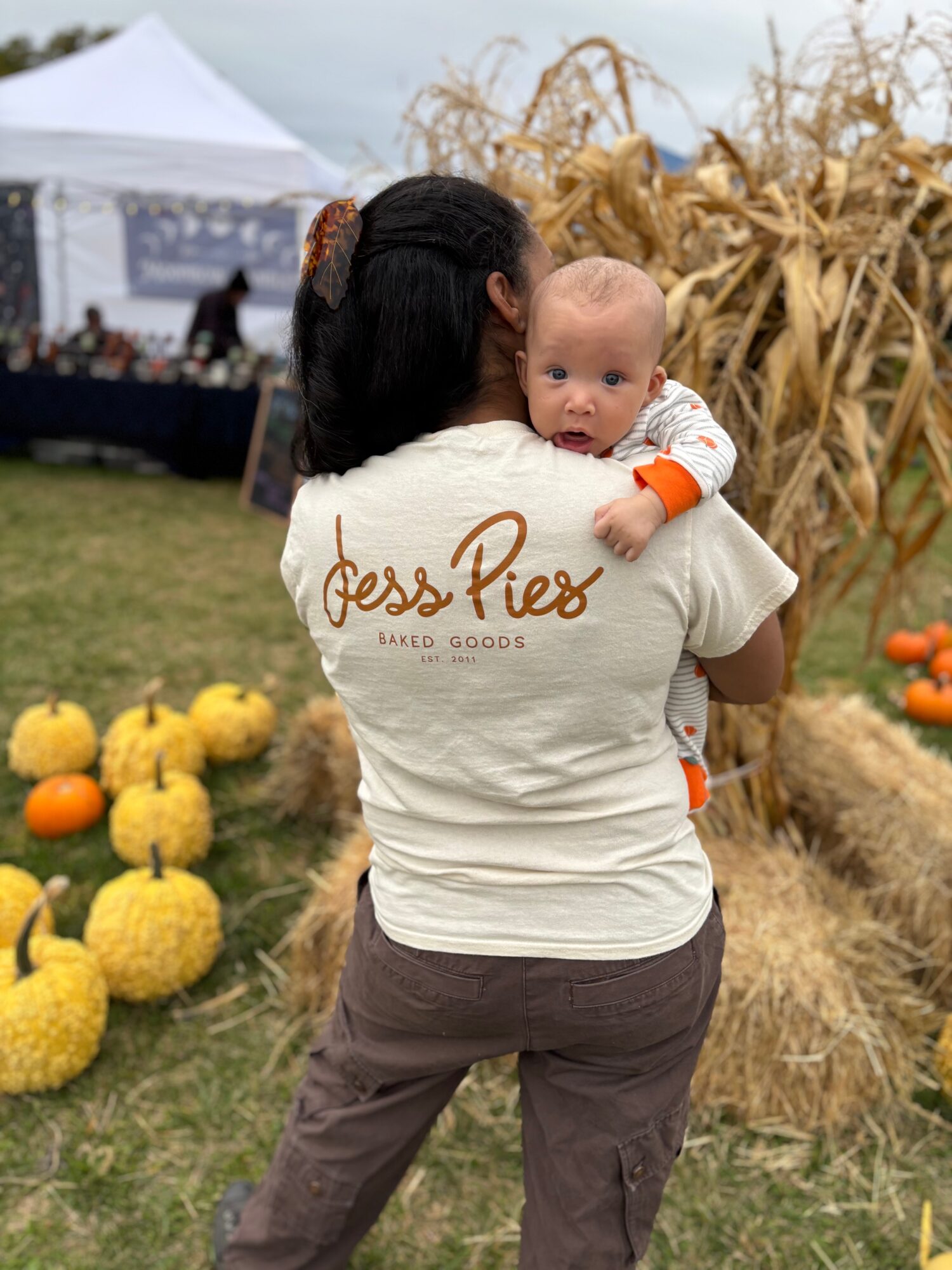 Person holding a baby beside pumpkins and dried cornstalks at an outdoor event.