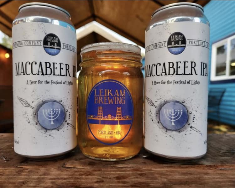 Two white beer cans flank an amber jar labeled Leikam Brewing on a wooden surface.