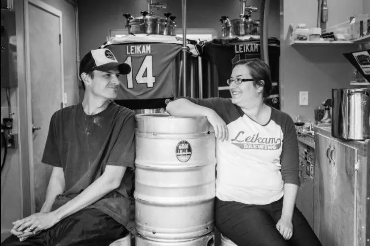 Two people sit beside a metal keg in a room with sports jerseys on the wall.