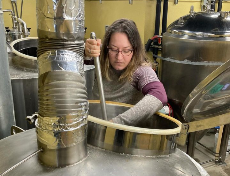 A person stirs a large stainless-steel brewing vessel beside tanks and metal pipes.