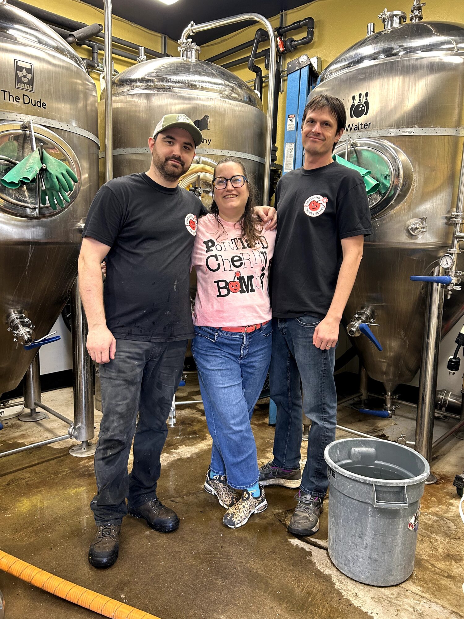 Three people stand between large stainless steel brewing tanks, with a bucket on the floor.
