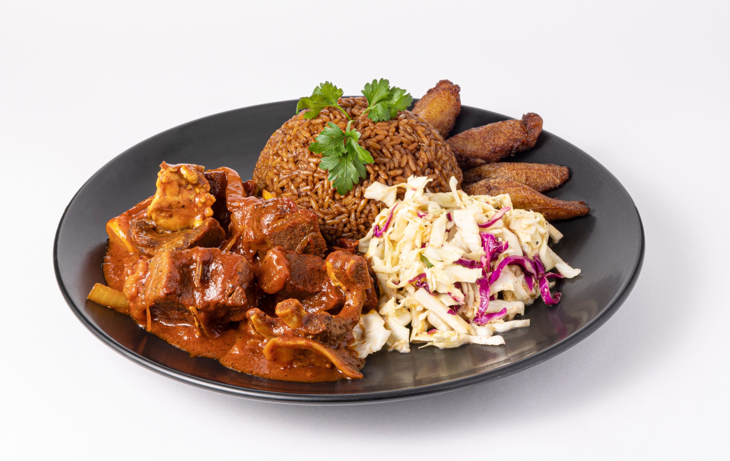 A plate of sauced meat, rice, fried plantains, and shredded cabbage salad garnished with herbs.