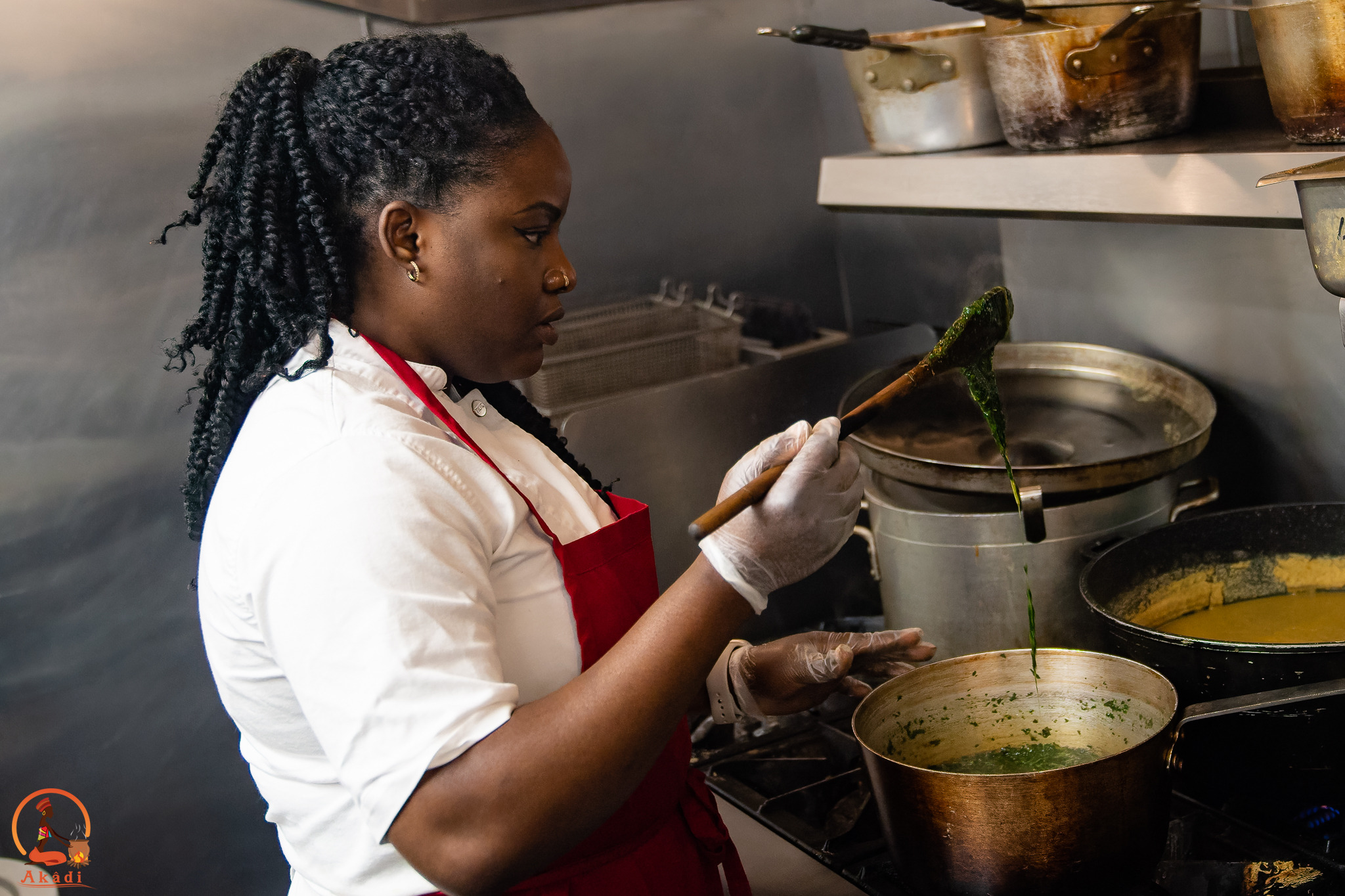 A person in a white shirt and red apron stirs a pot on a commercial stovetop.