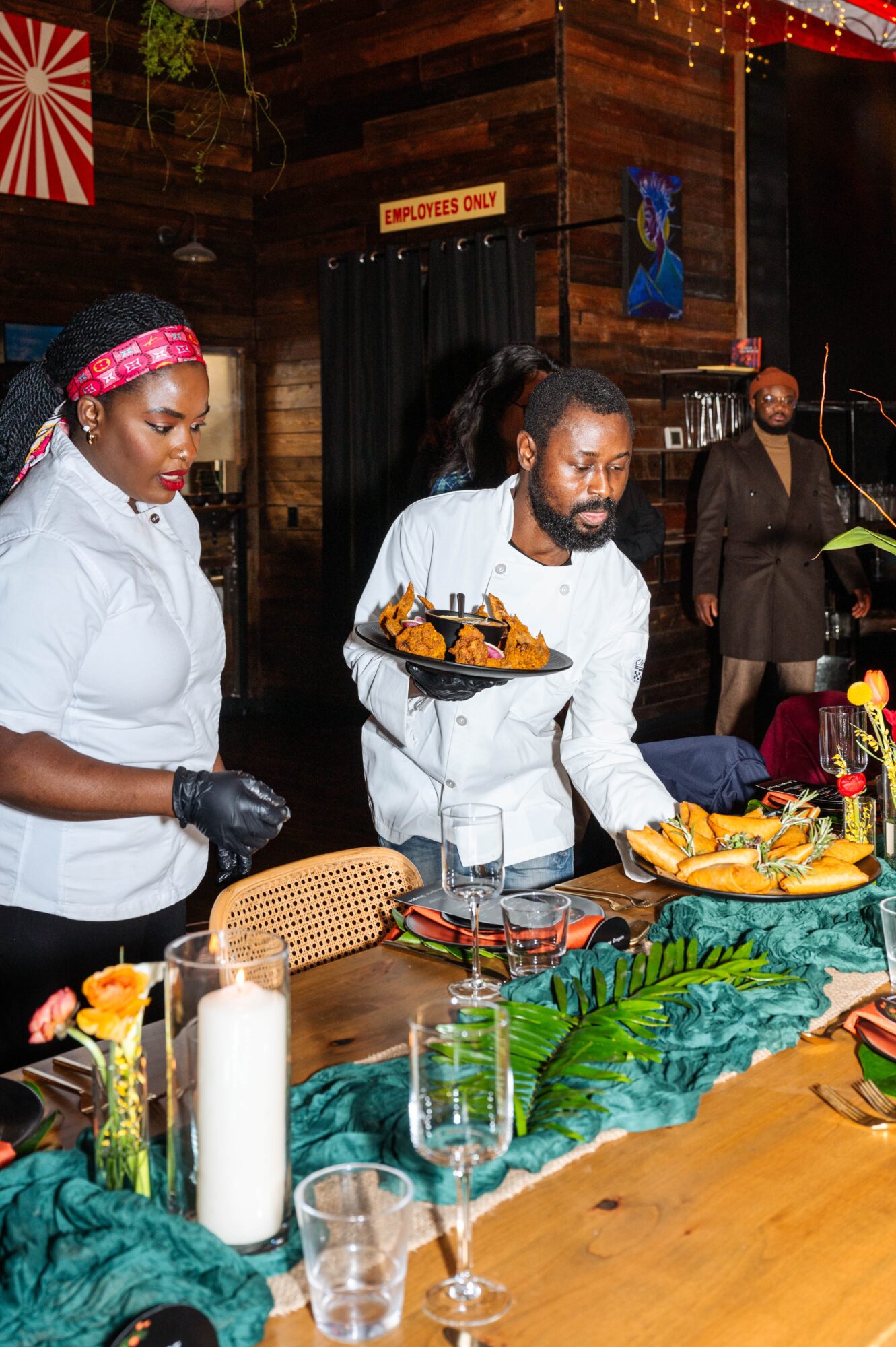 Two people serve food at a buffet table decorated with greenery, glasses, and dishes; another person stands in the background.