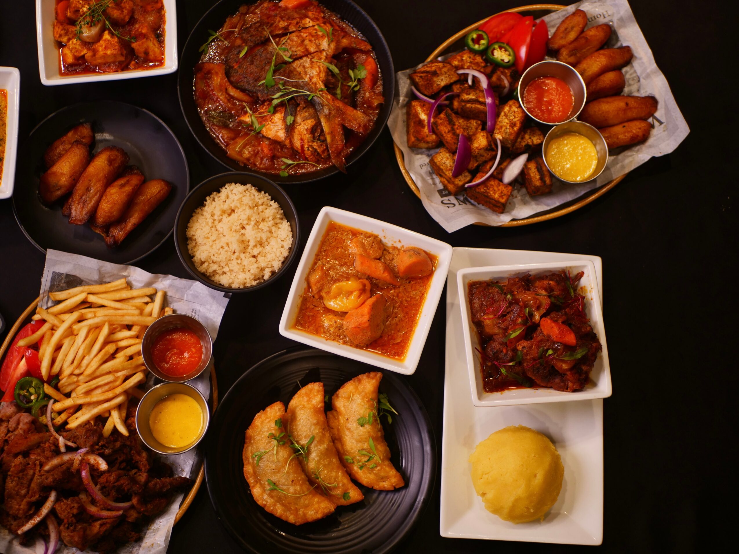 Overhead spread of assorted fried foods, fries, rice, stews, sauces, and sliced vegetables on a dark table.