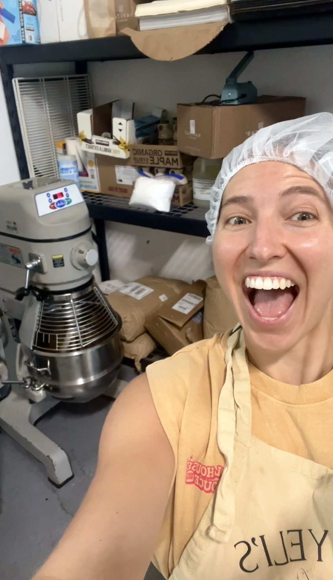 Person wearing a hairnet and apron smiles beside a large mixer and stacked boxes in a workroom.