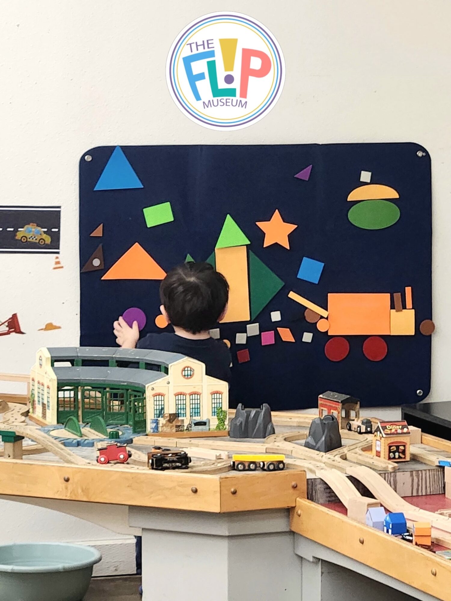 A child stands at a wooden train table beneath a colorful geometric wall display and museum logo.