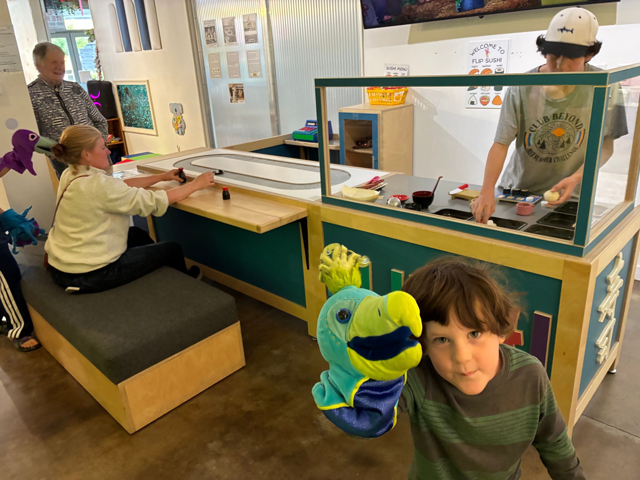People stand beside a colorful plush parrot in a room with display cases and a wooden table.