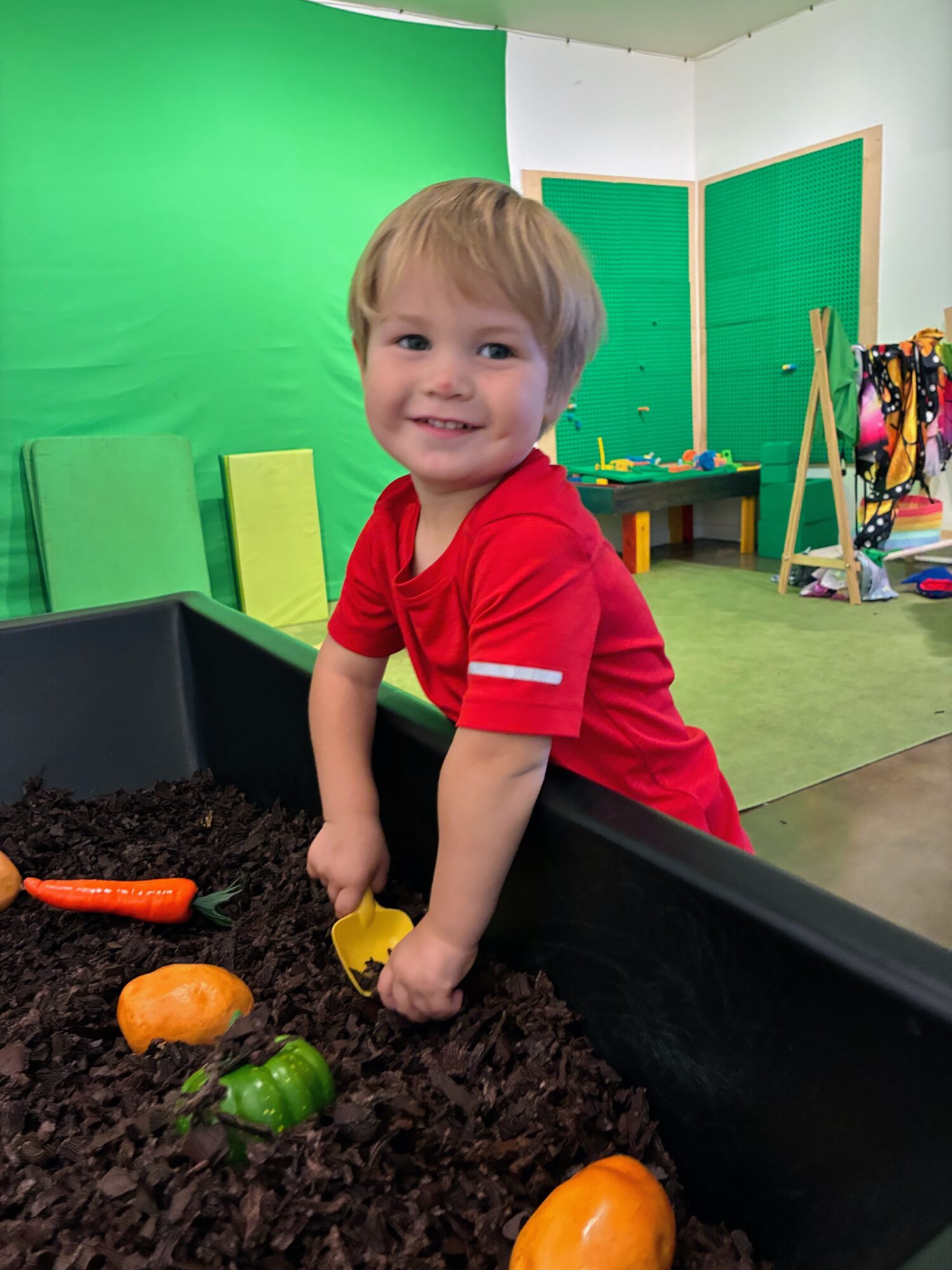 A young child in a red shirt plays with toy vegetables in a soil-filled indoor planter.