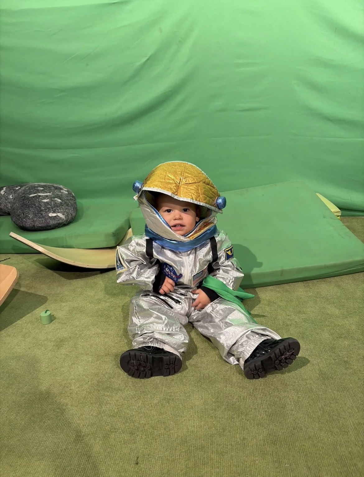 A seated child wears a yellow helmet and silver protective suit on a green floor in front of a green backdrop.