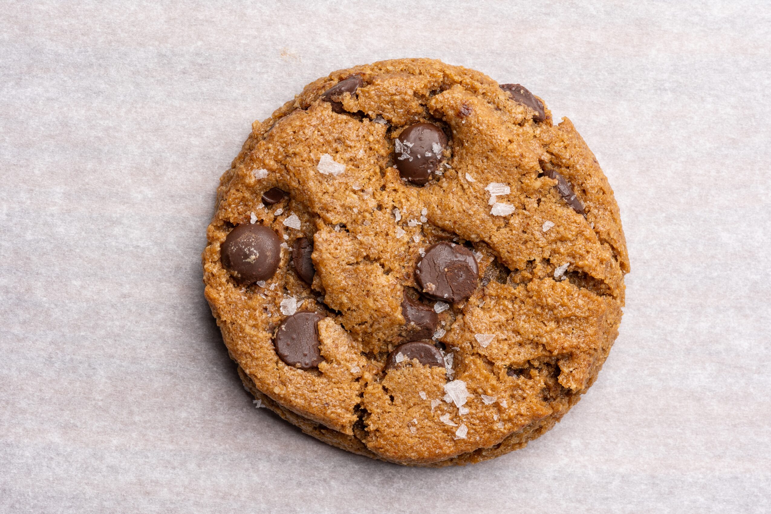 Round chocolate chip cookie with flaky salt on a light surface.