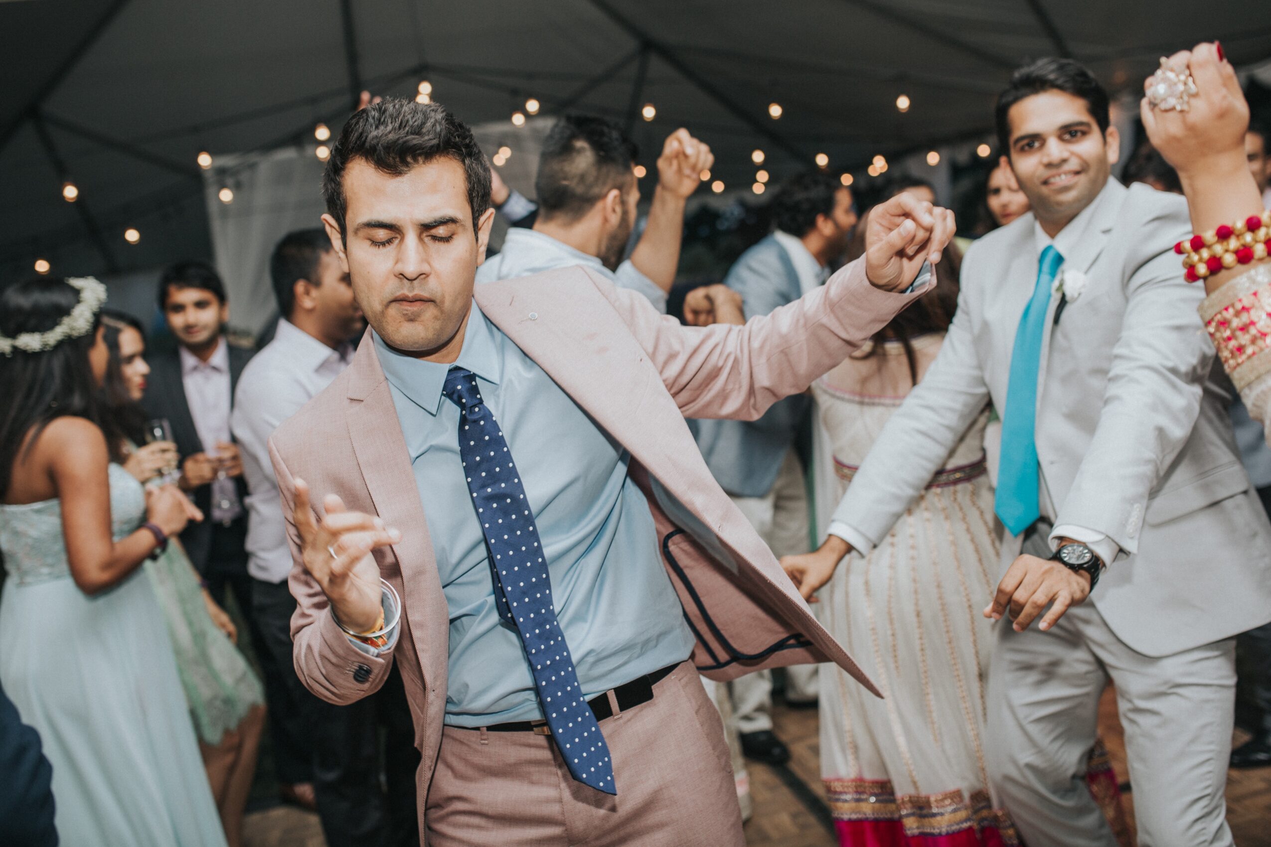 People dance beneath a tent decorated with string lights; a man in a pink suit moves in the foreground.