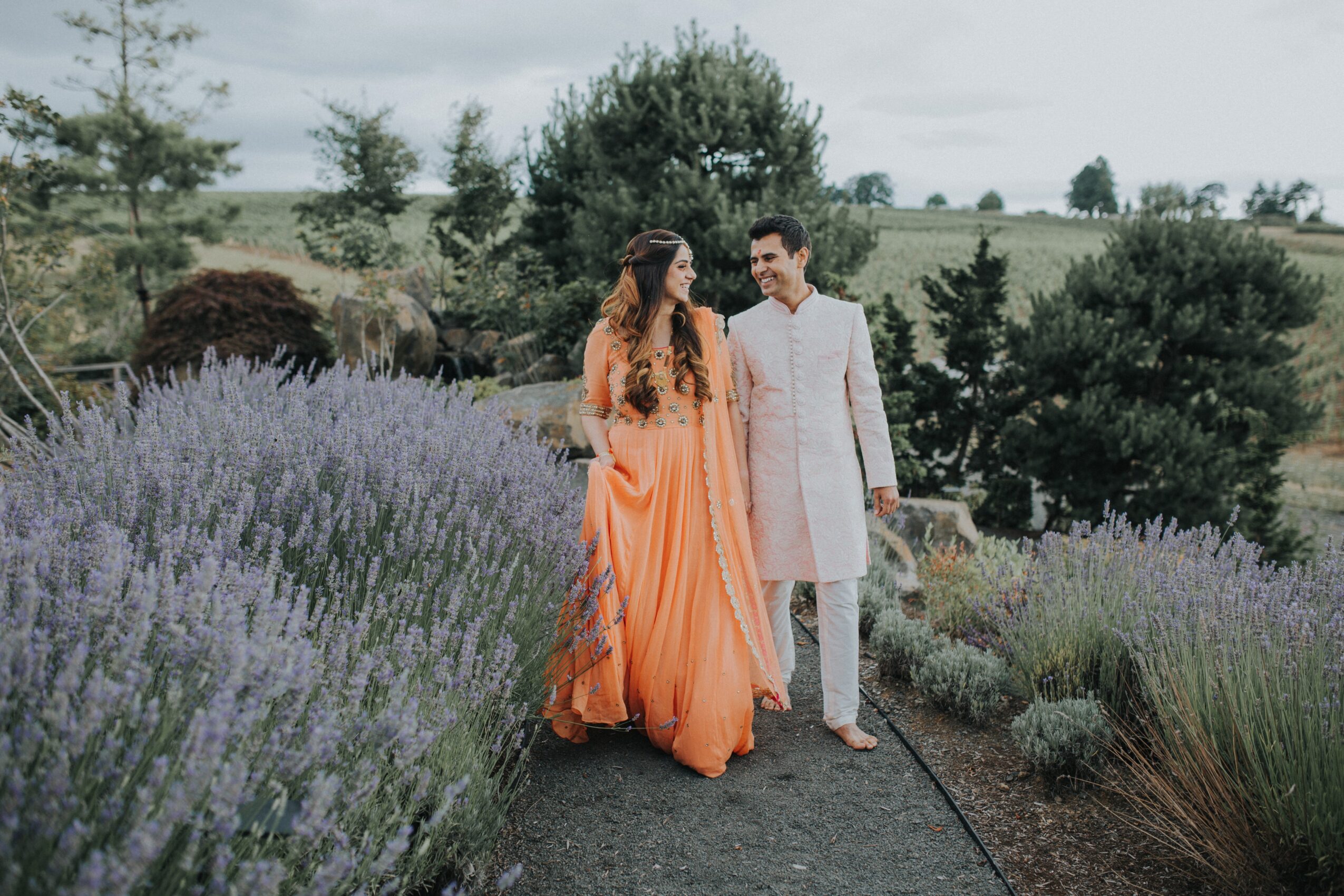 Two people stand together on a garden path among flowering plants and trees.