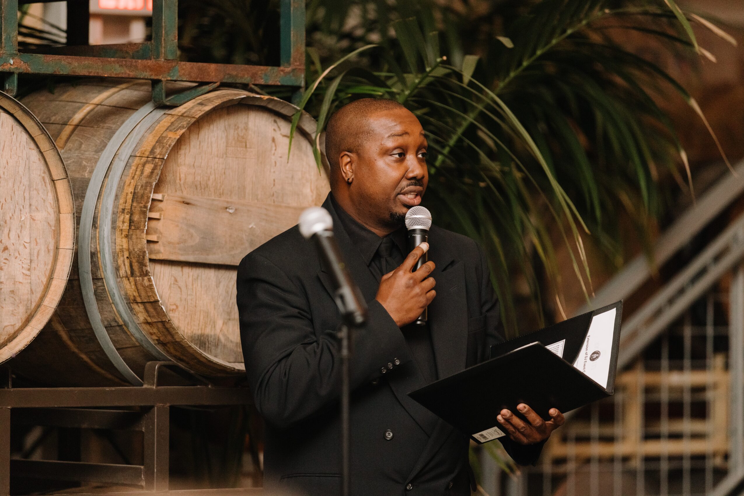 A man speaks into a microphone while holding an open folder in front of large wooden barrels.