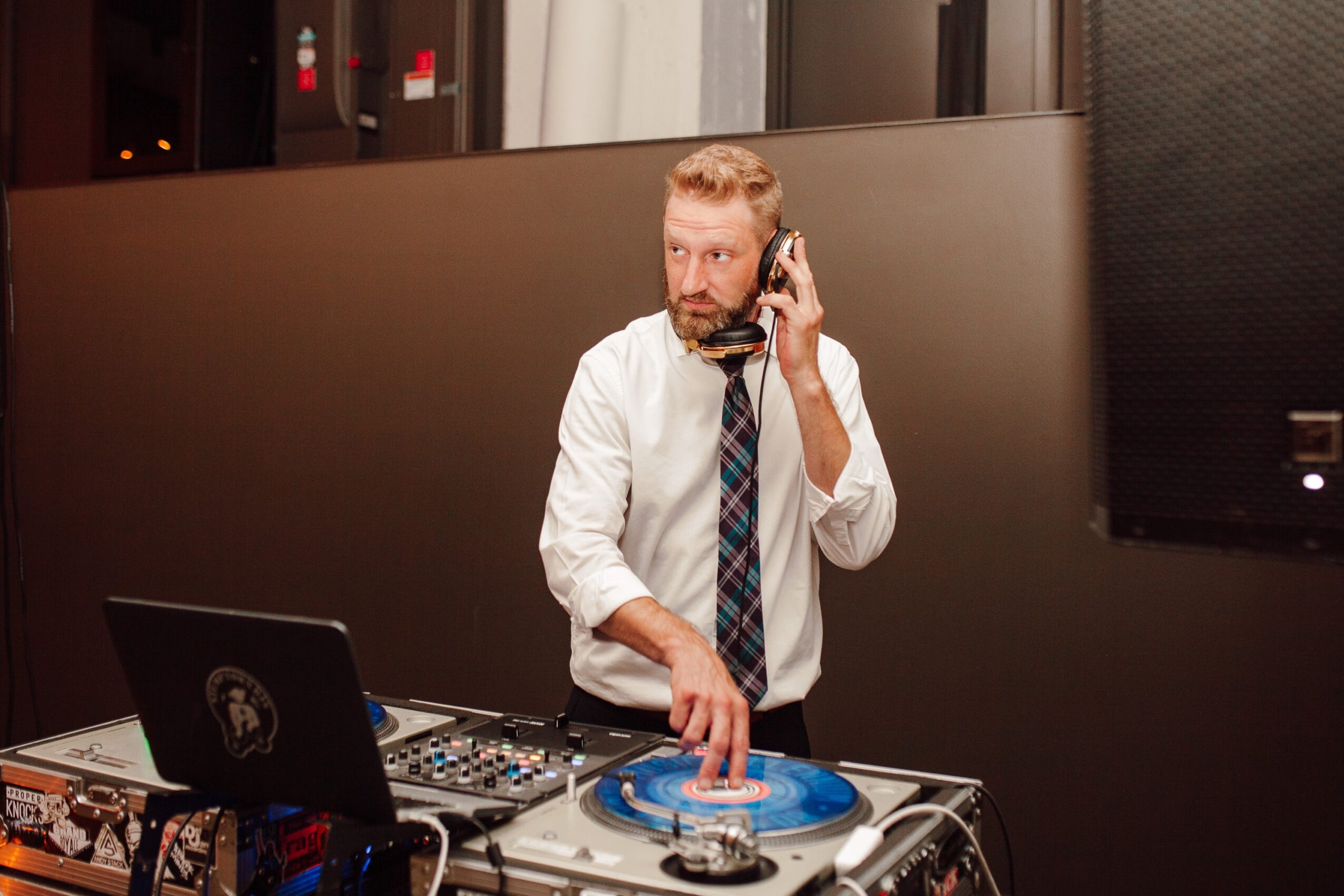 A person wearing a white shirt and patterned tie talks on a phone while operating a turntable and laptop.