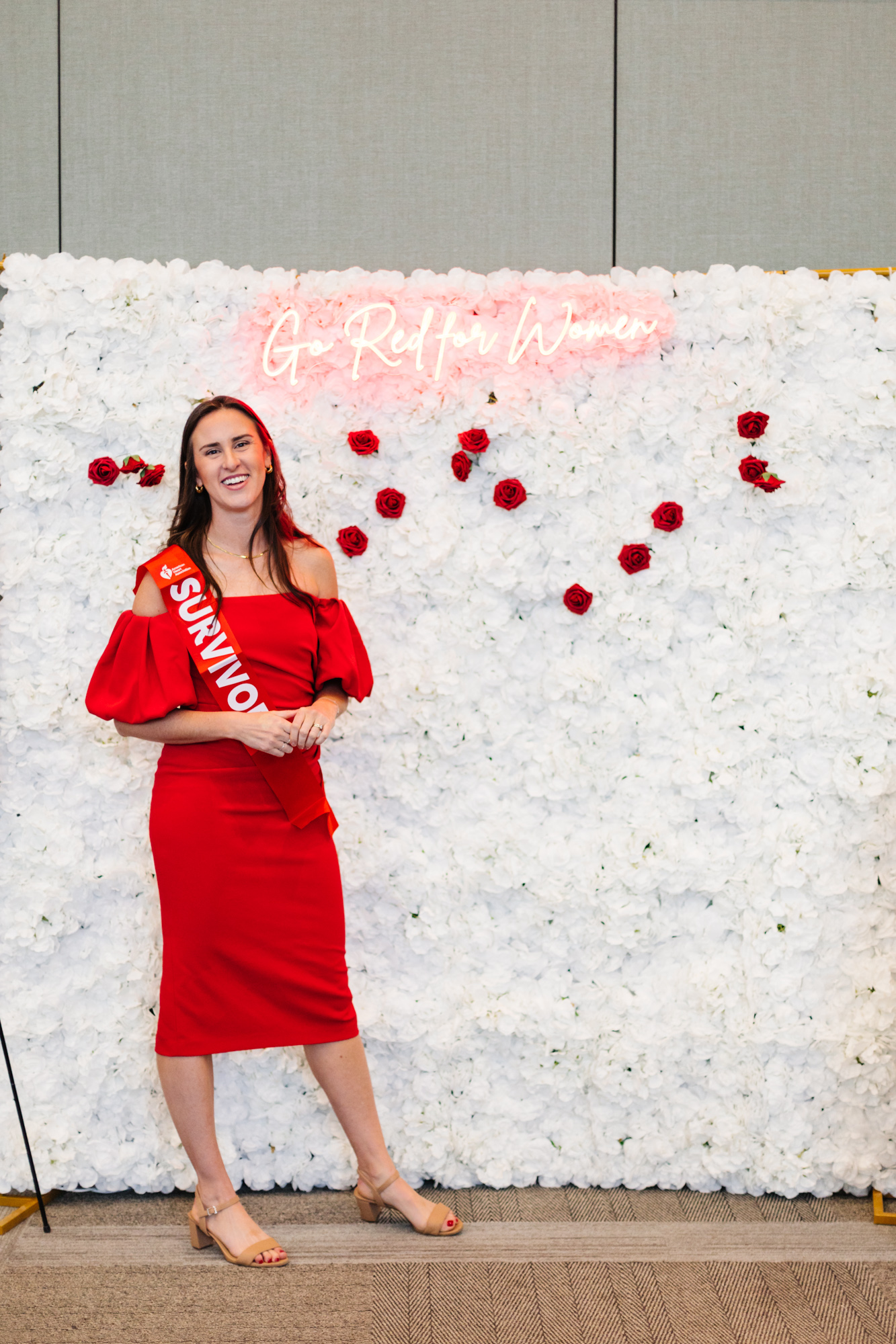 A person in a red dress stands before a white flower wall with red roses and a glowing sign.