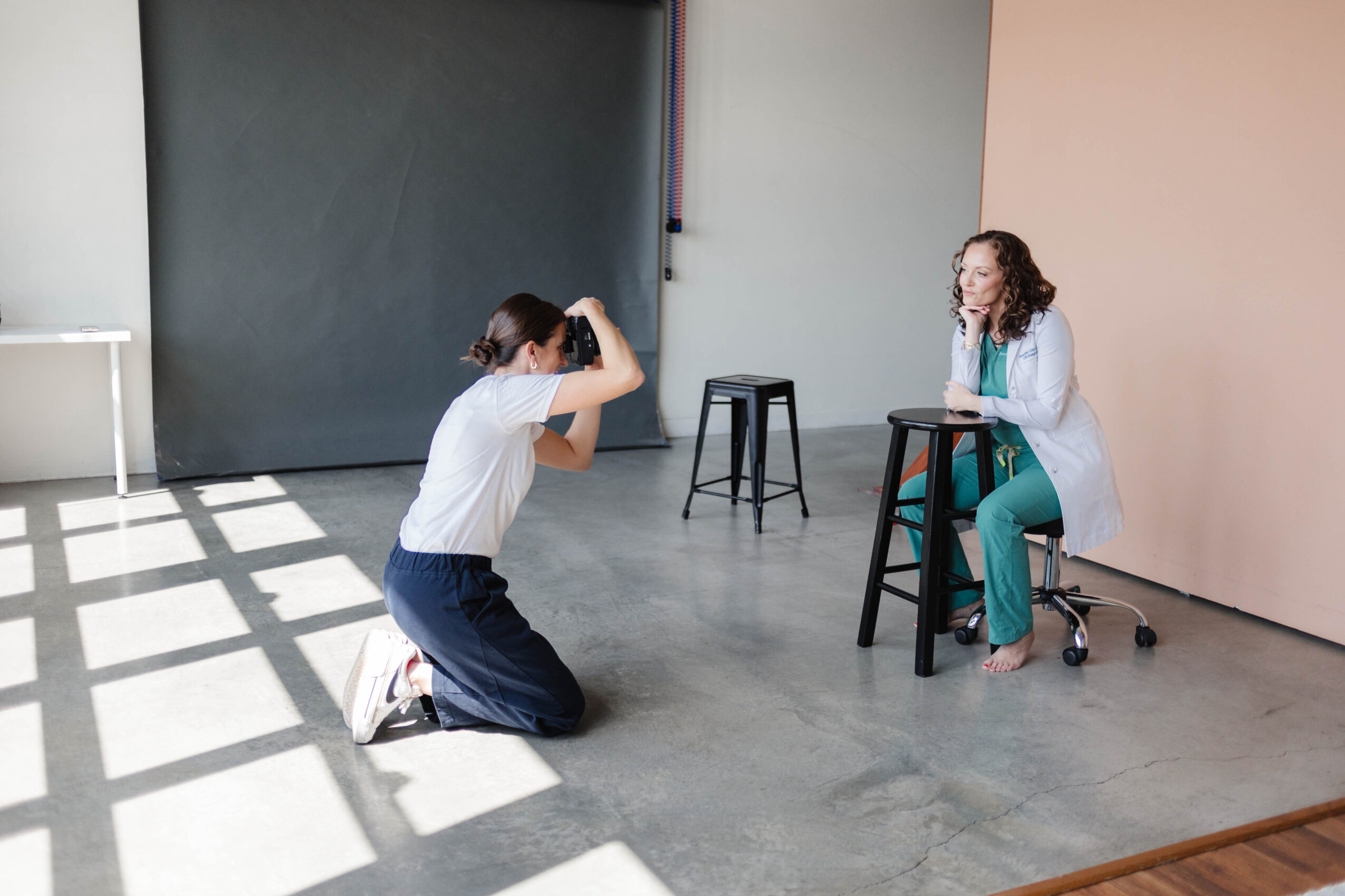 A kneeling person photographs a seated person beside a stool in a bright studio.