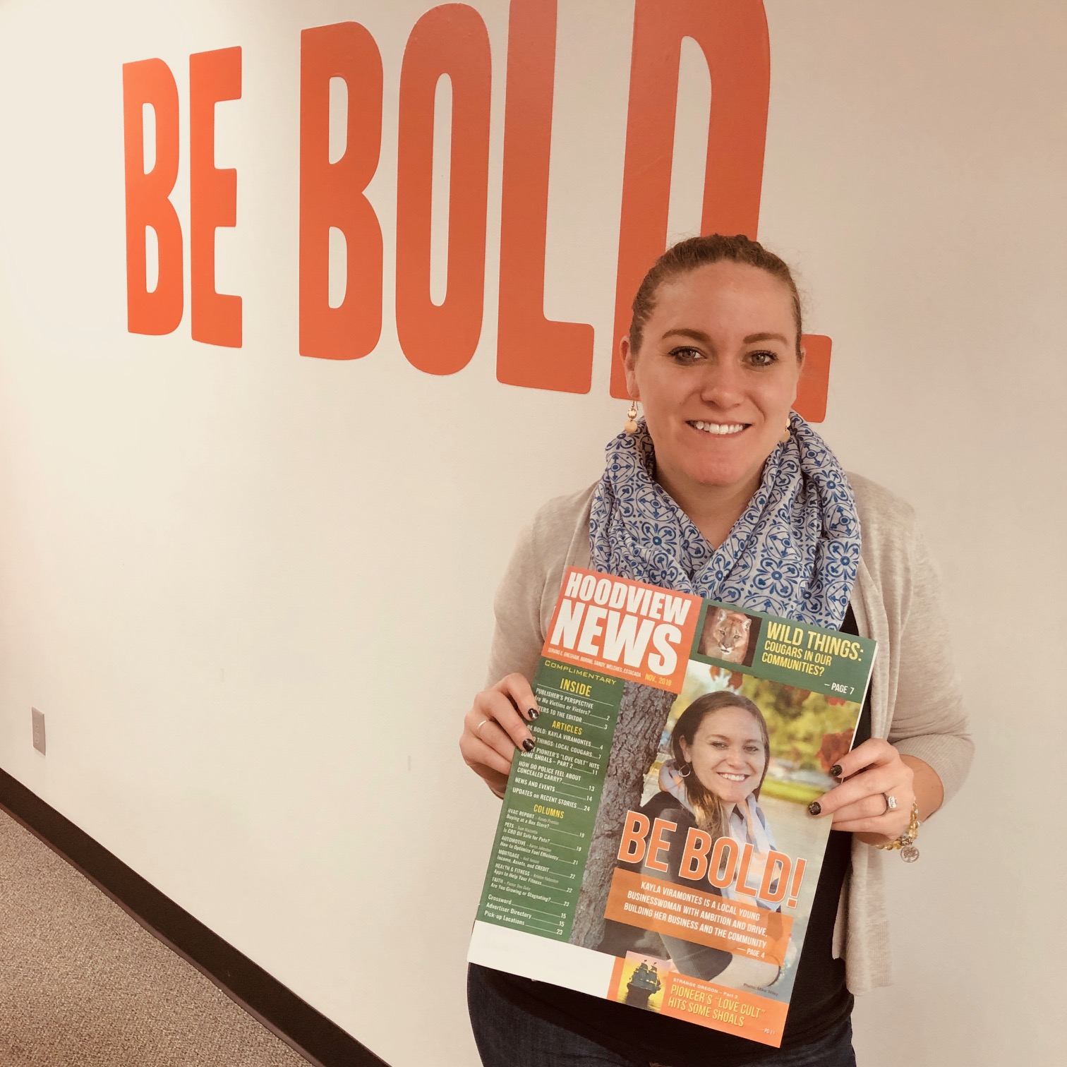 A smiling person holds a magazine in front of a wall with large orange lettering.