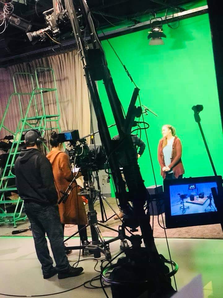 A camera crew films a person standing before a green screen on a studio set.
