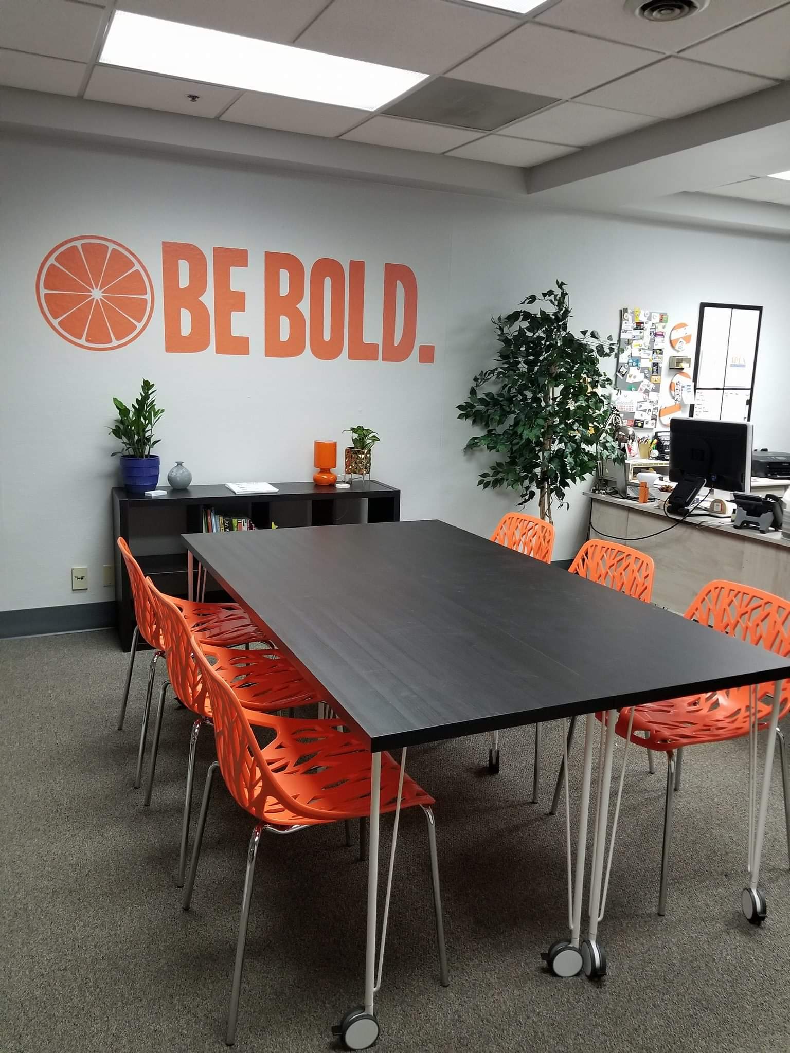 Conference room with a long dark table, orange chairs, a potted tree, and an orange “BE BOLD.” wall graphic.