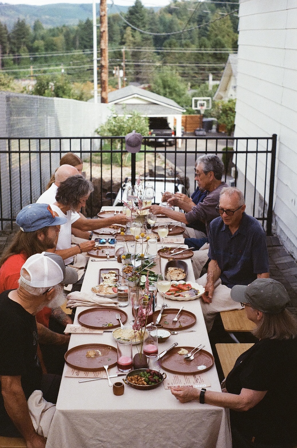 People sit around a long outdoor dining table set with plates, glasses, and food.