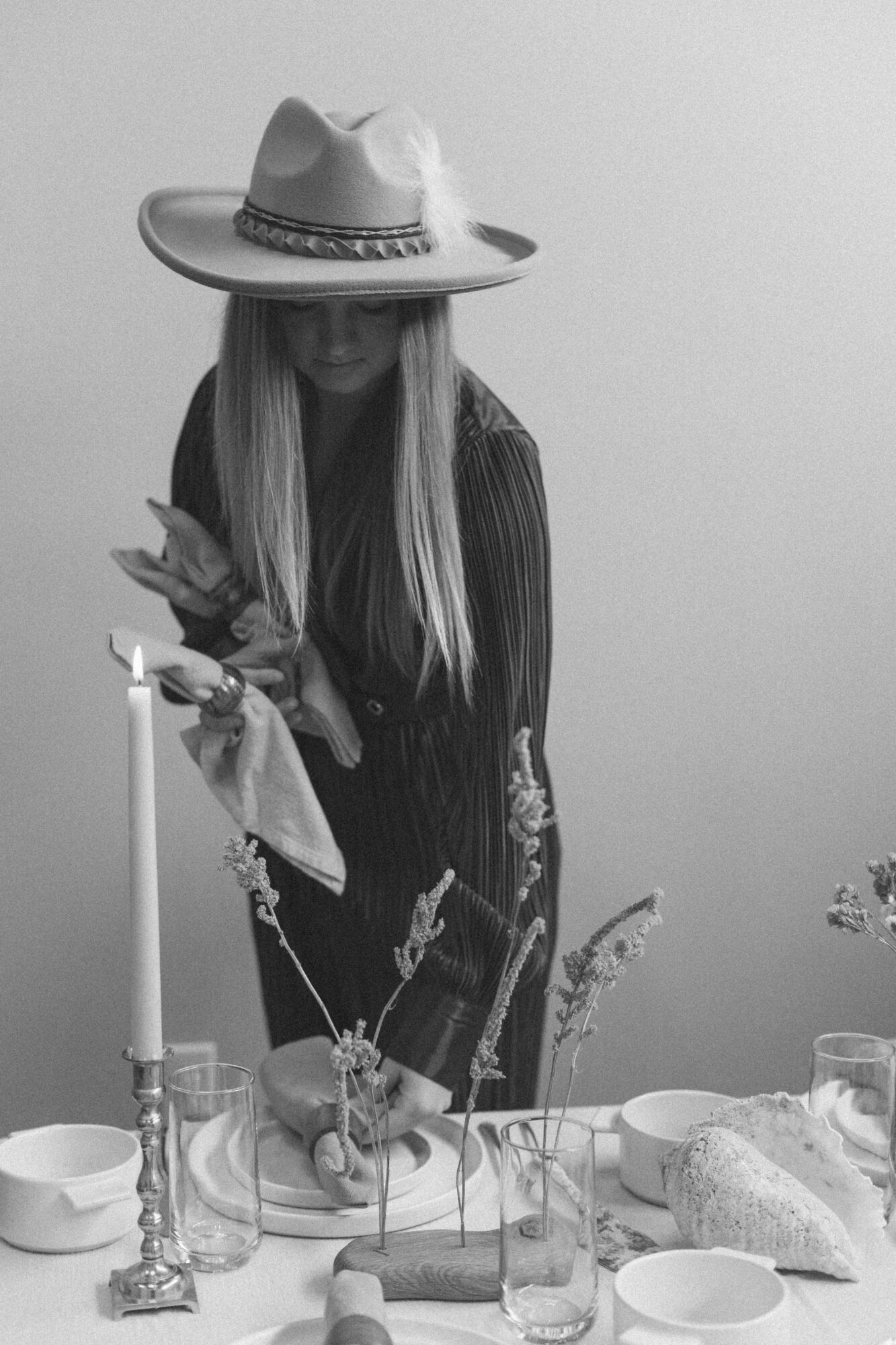Person wearing a wide-brimmed hat stands behind a table with plates, glasses, candles, and flowers.