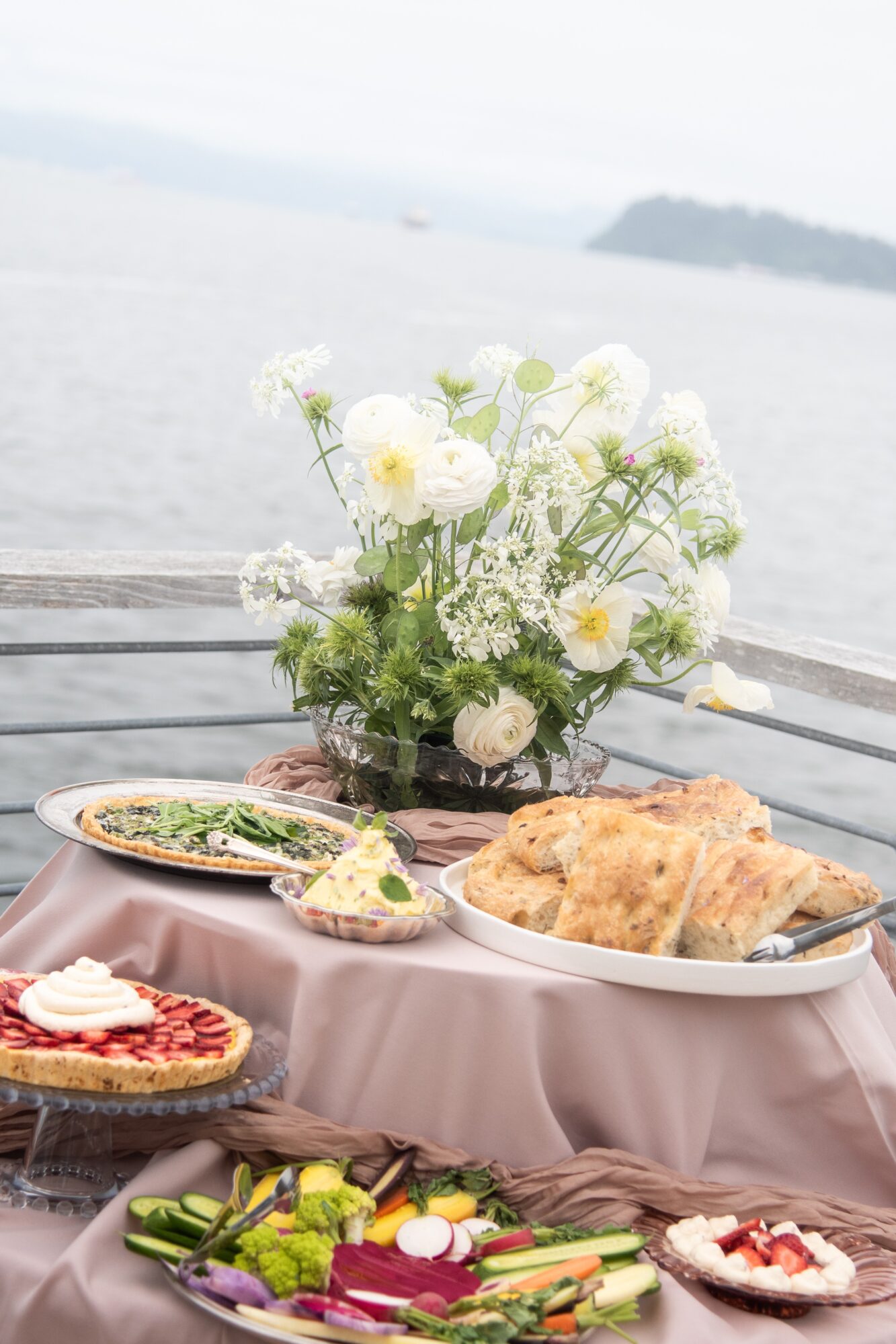 A table with pastries, plates of food, and a vase of white flowers beside a waterfront.