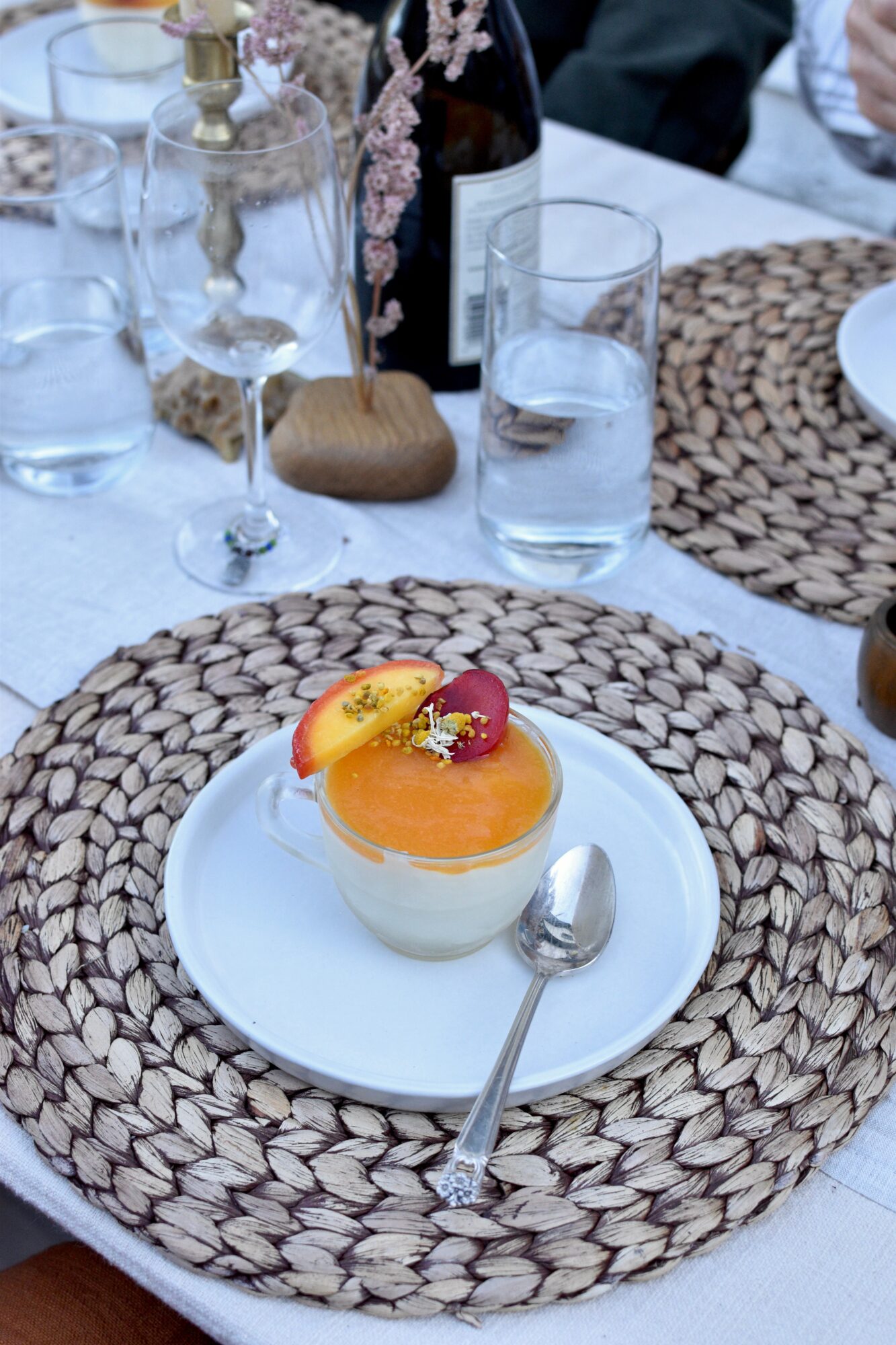 A small orange-topped dessert sits on a white plate over a woven placemat, beside glasses and a spoon.